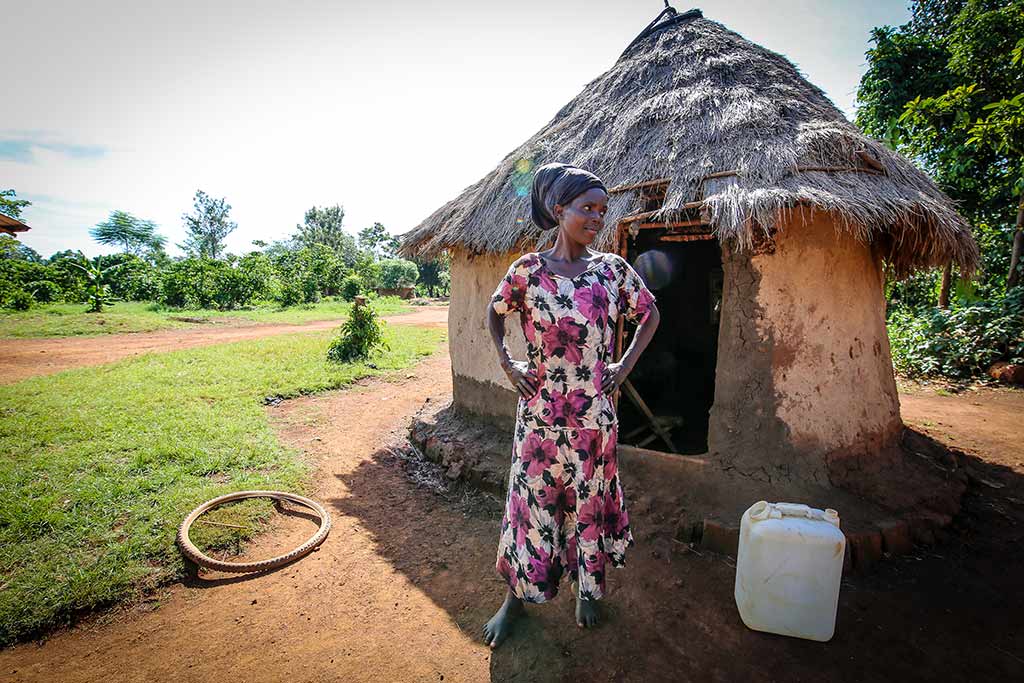 A woman in a long dress stands outside of her home made of thatched roof and mud walls.